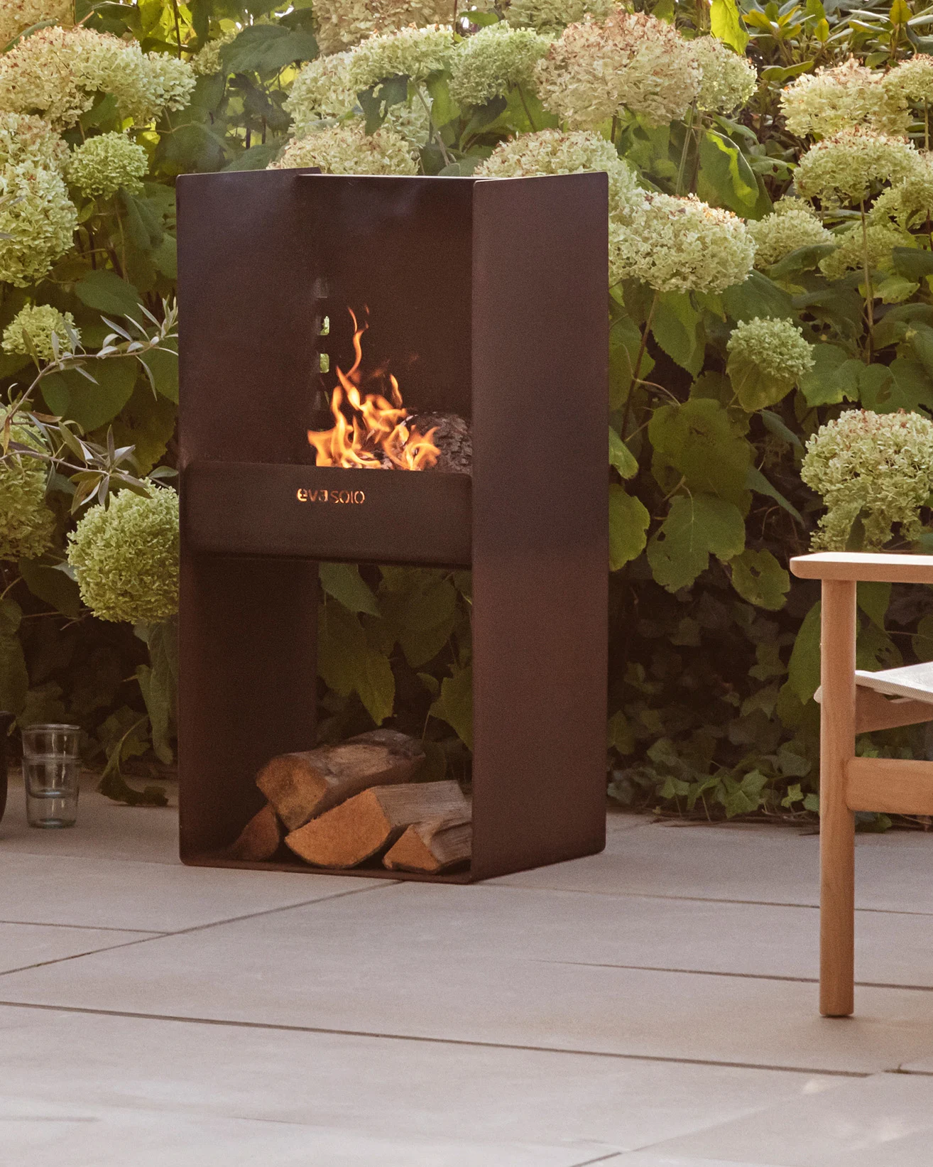 Moderner dunkler Metall-Außenkamin mit brennendem Feuer, gestapelten Holzscheiten darunter, umgeben von grünen Pflanzen und einer Terrasse.