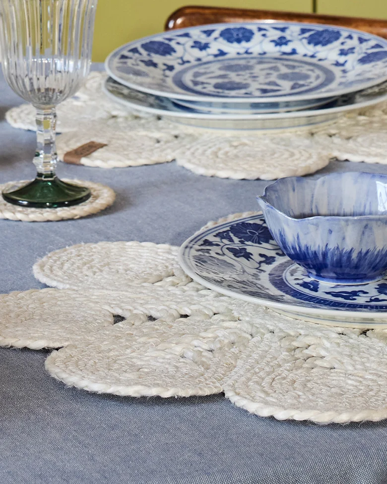 Gedeckter Tisch mit blau-weißem Geschirr, weißen geflochtenen Blumenplatzsets und einem gerippten Glas.
