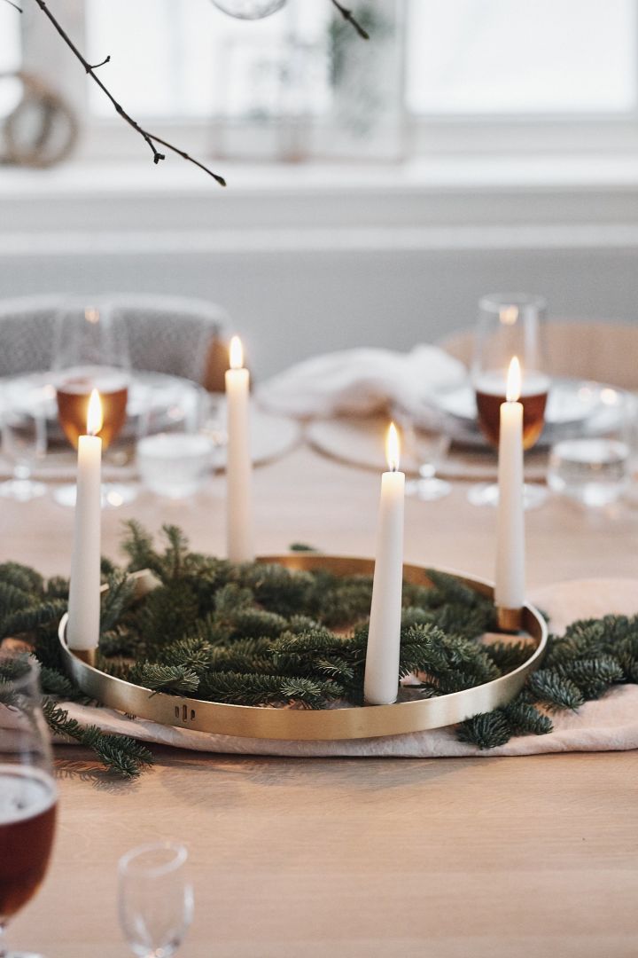 Fichtenzweige und ein runder Kerzenhalter aus Messing mit weißen Kerzen stehen in der Mitte eines weißen Weihnachtstischs und verbreiten eine festliche Stimmung.