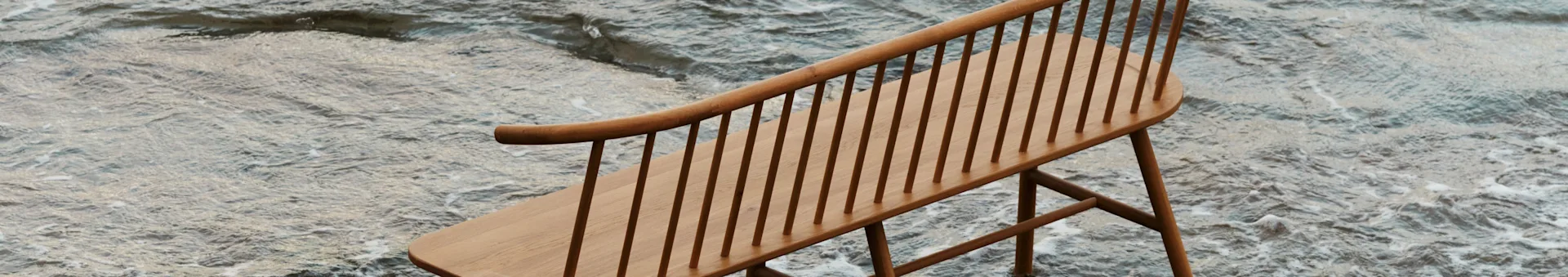 Eine helle Holzbank mit Spindelrücken und gebogener Rückenlehne, teilweise in seichtem, gekräuseltem Wasser versenkt.