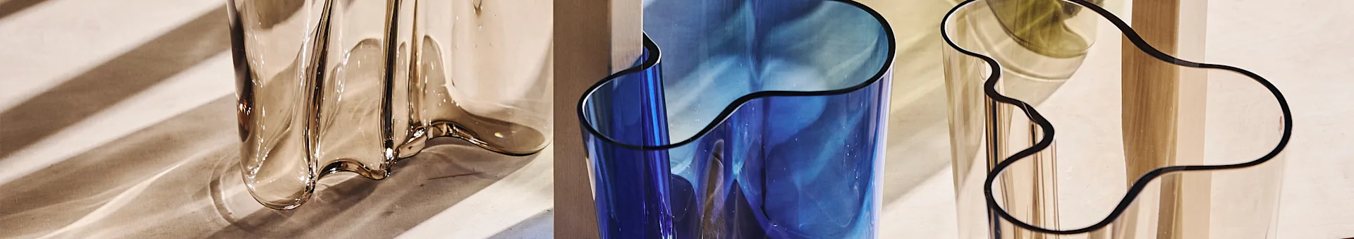 Nahaufnahme von drei wellenförmigen, durchscheinenden Vasen: bernsteinfarben, blau und klar, die gestreifte Schatten reflektieren.