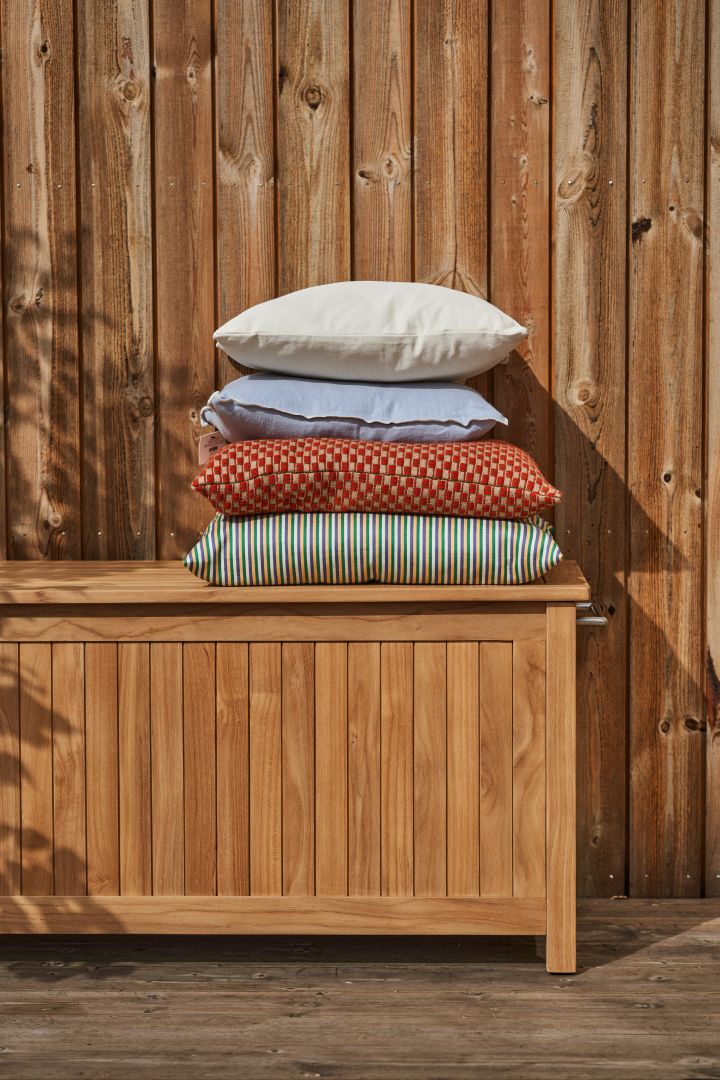 Bringen Sie Leben, Farbe und Komfort auf Ihre Terrasse mit Kissen in verschiedenen Farben, Mustern und Materialien, hier Kissen in Beige, Hellblau, Rot und Grün auf einer Teakholz-Kissenbox.