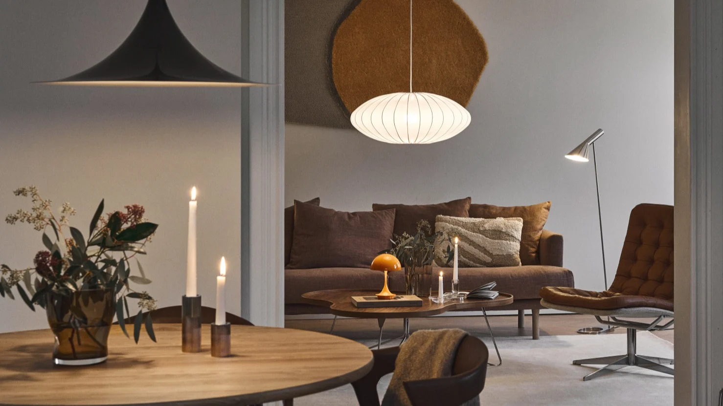 Ein warm beleuchtetes Ess- und Wohnzimmer mit Designerlampen, braunem Sofa und Ledersessel.