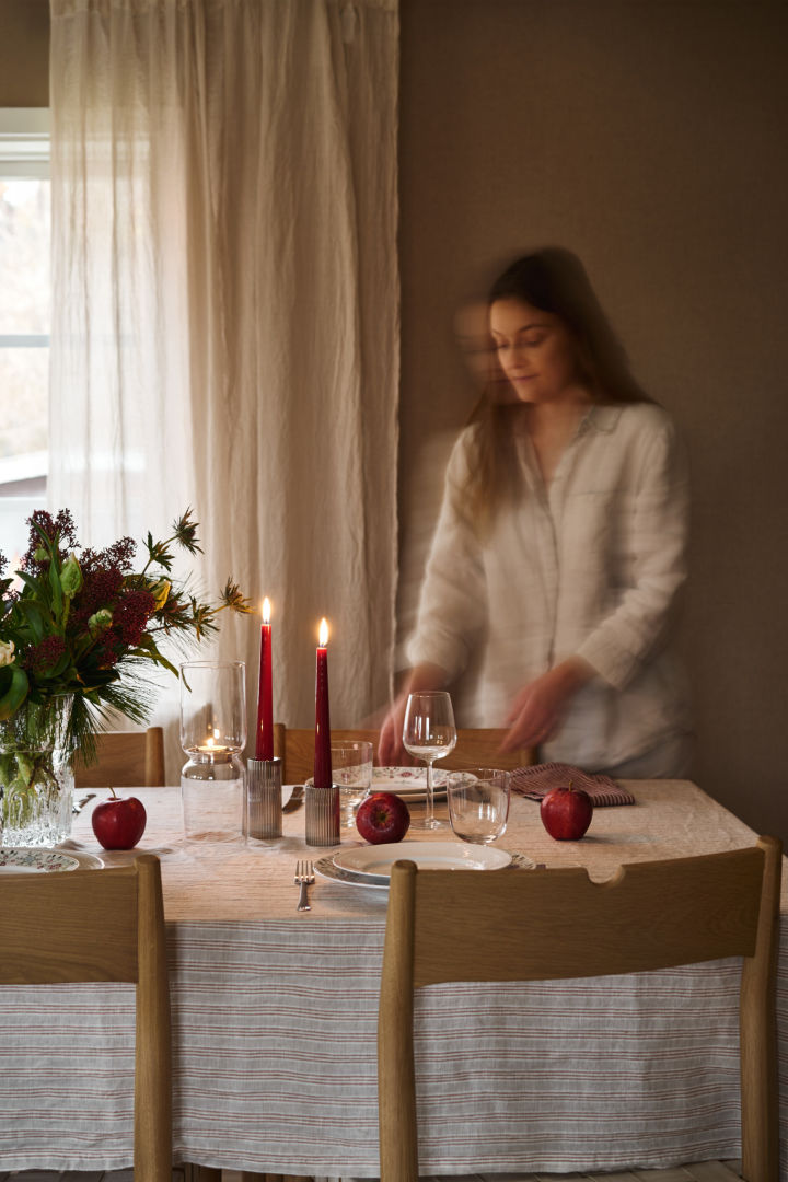 Eine Frau deckt einen einfachen Weihnachtstisch mit roten Kerzen, gemustertem Weihnachtsporzellan und einem einfachen gestreiften Tischtuch. 