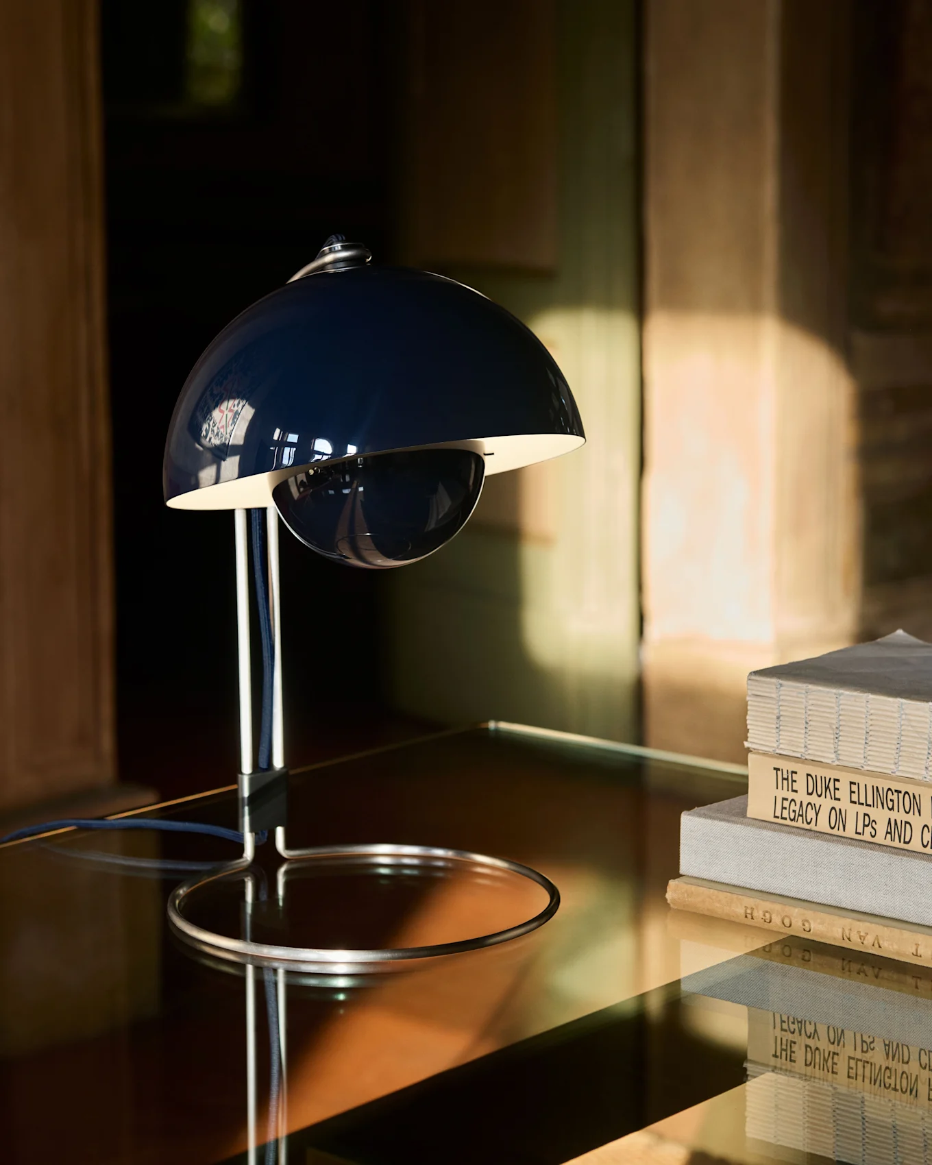 Eine dunkelblaue, glockenförmige Tischlampe mit Chromfuß steht auf einem reflektierenden Tisch neben einem Stapel Bücher.
