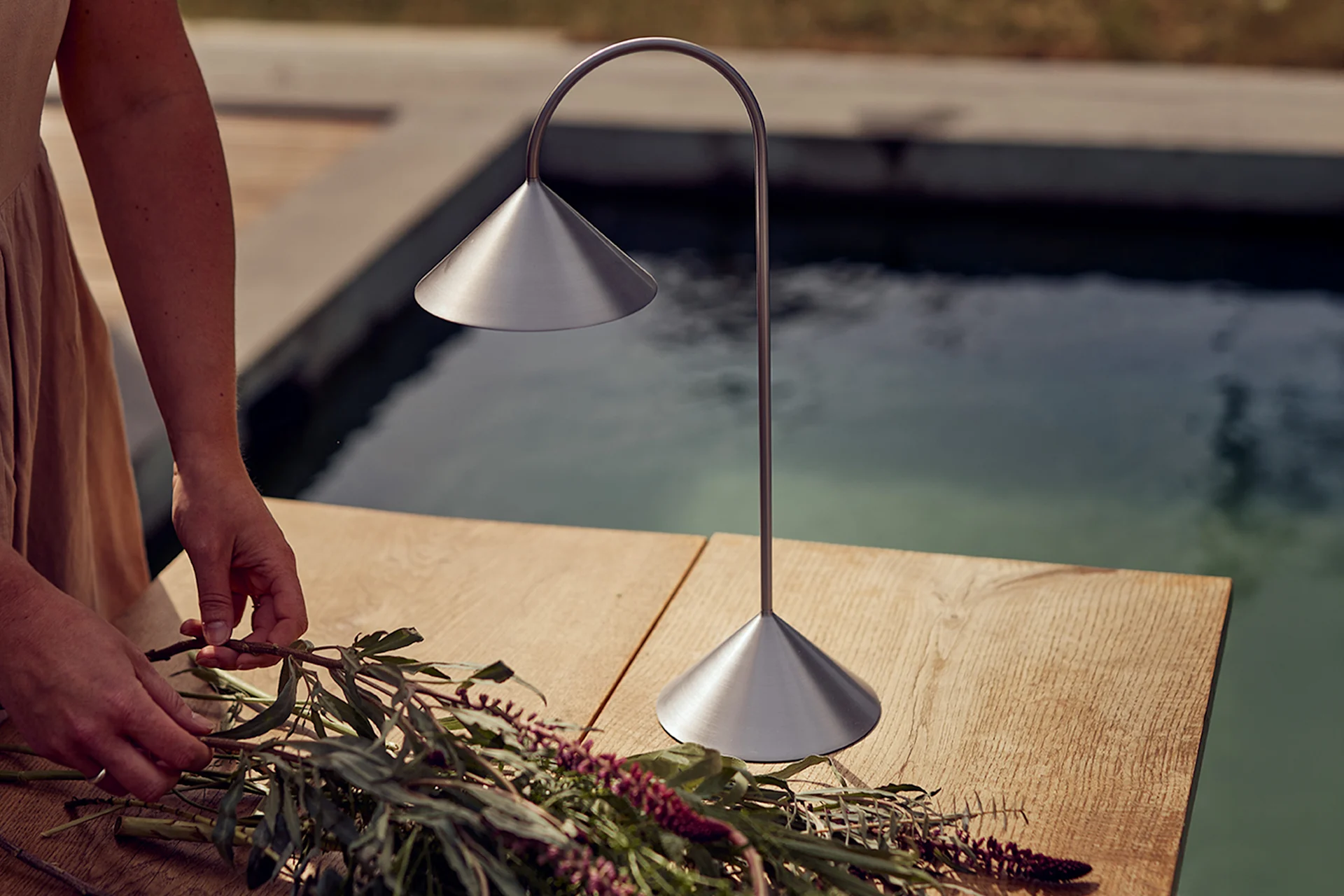 Hände arrangieren Blumen auf einem Holztisch neben einer gebürsteten Metall-Tischlampe am Pool.