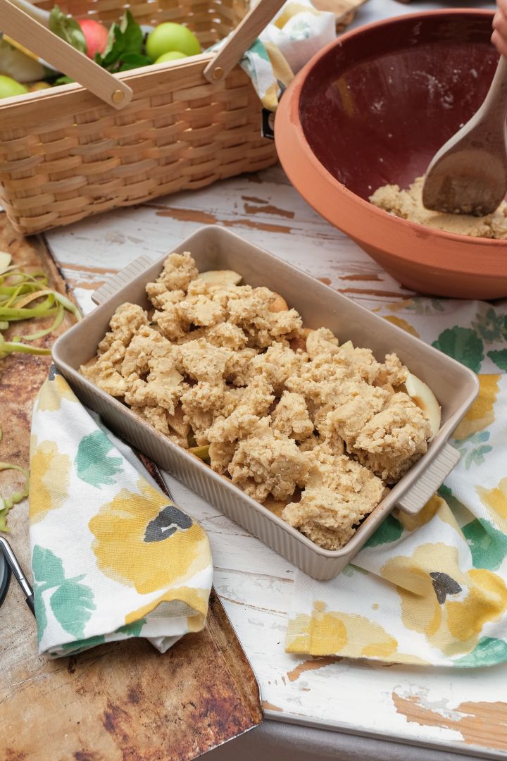 Das Bild zeigt die Flora-Backform von Sagaform, eine beigefarbene Auflaufform mit Henkel, in der ein ungebackener Apfelkuchen liegt.