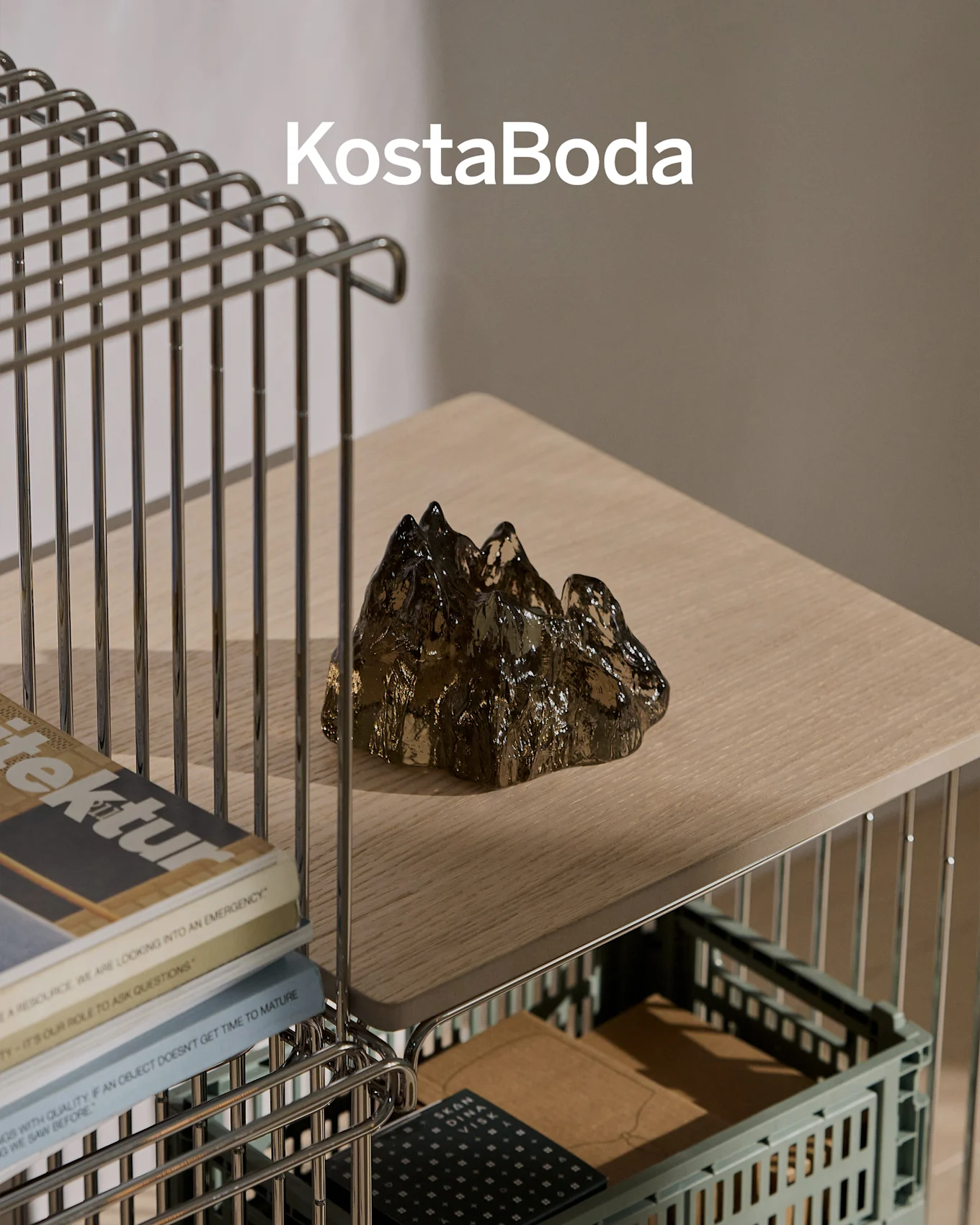 Markenname KostaBoda über einer dunklen, transparenten, bergförmigen Glasskulptur auf einem hellen Holzregal.