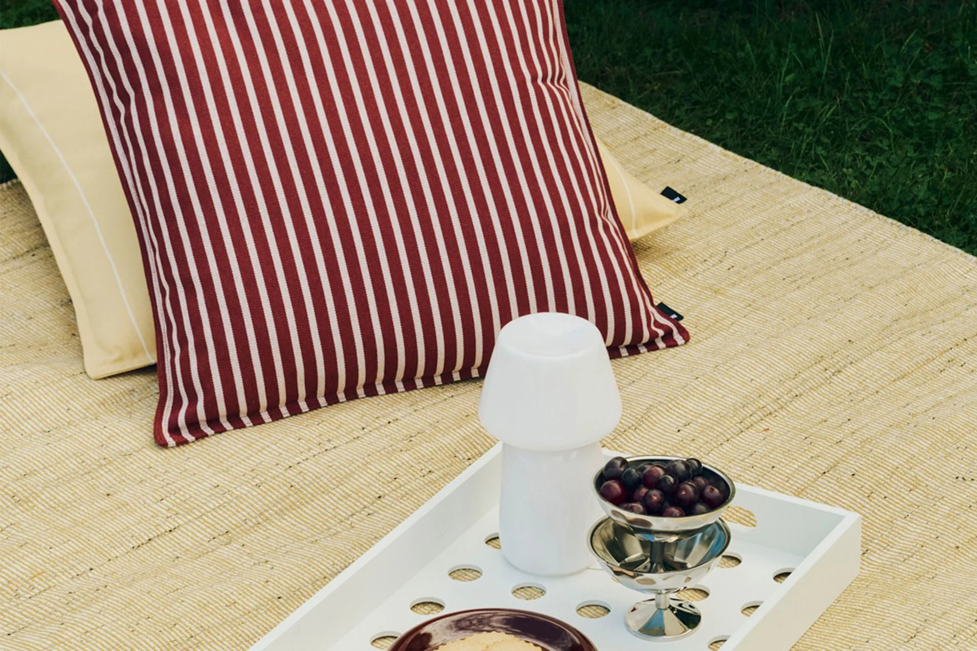 Outdoor-Picknick-Setup mit gestreiftem rot-weißem Kissen, gelbem Kissen, weißem Tablett mit Pilzlampe und einer Schüssel Beeren.