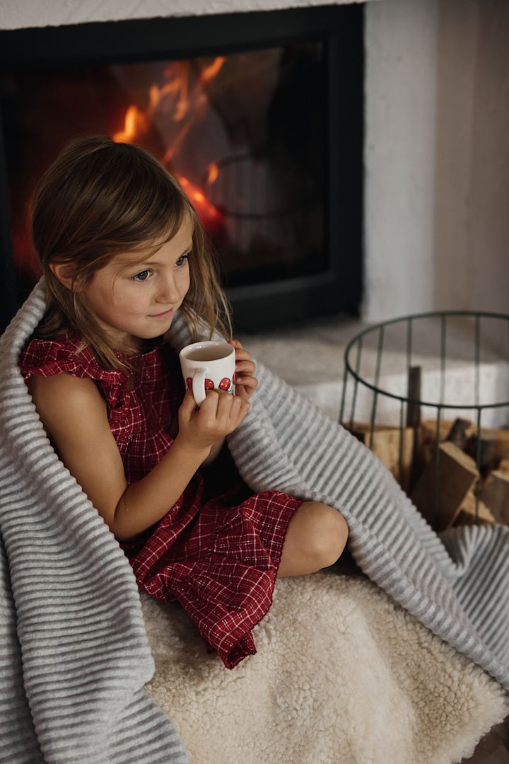 Zum schwedischen Weihnachtsfest gehört auch eine Tasse heißer Schokolade oder Glögg. Hier sehen Sie ein Mädchen mit der Children of the forest Tasse und der umgelegten Pleece Decke, beides von Design House Stockholm auf einem Sofa vor dem Kamin sitzen.