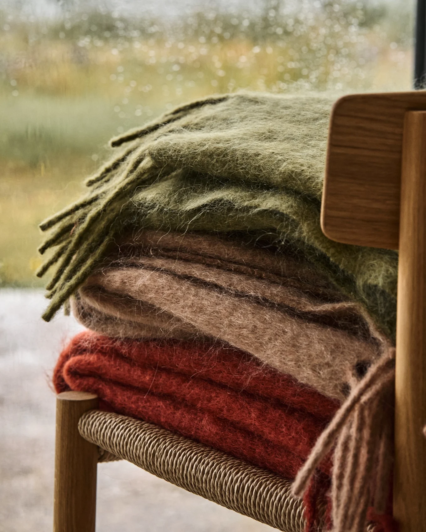 Stapel aus drei weichen Decken in Olivgrün, Beige und Rot-Orange auf einem Holzstuhl, mit regnerischem Fenster im Hintergrund.