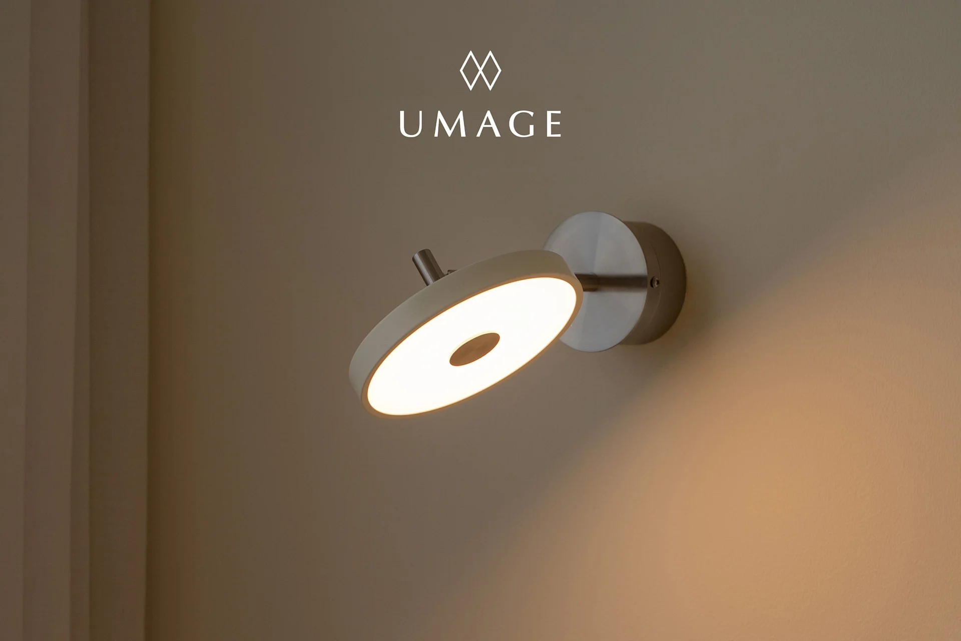Eine moderne, runde Wandleuchte mit warmem Licht, an einer hellbraunen Wand montiert. UMA-Logo oben.