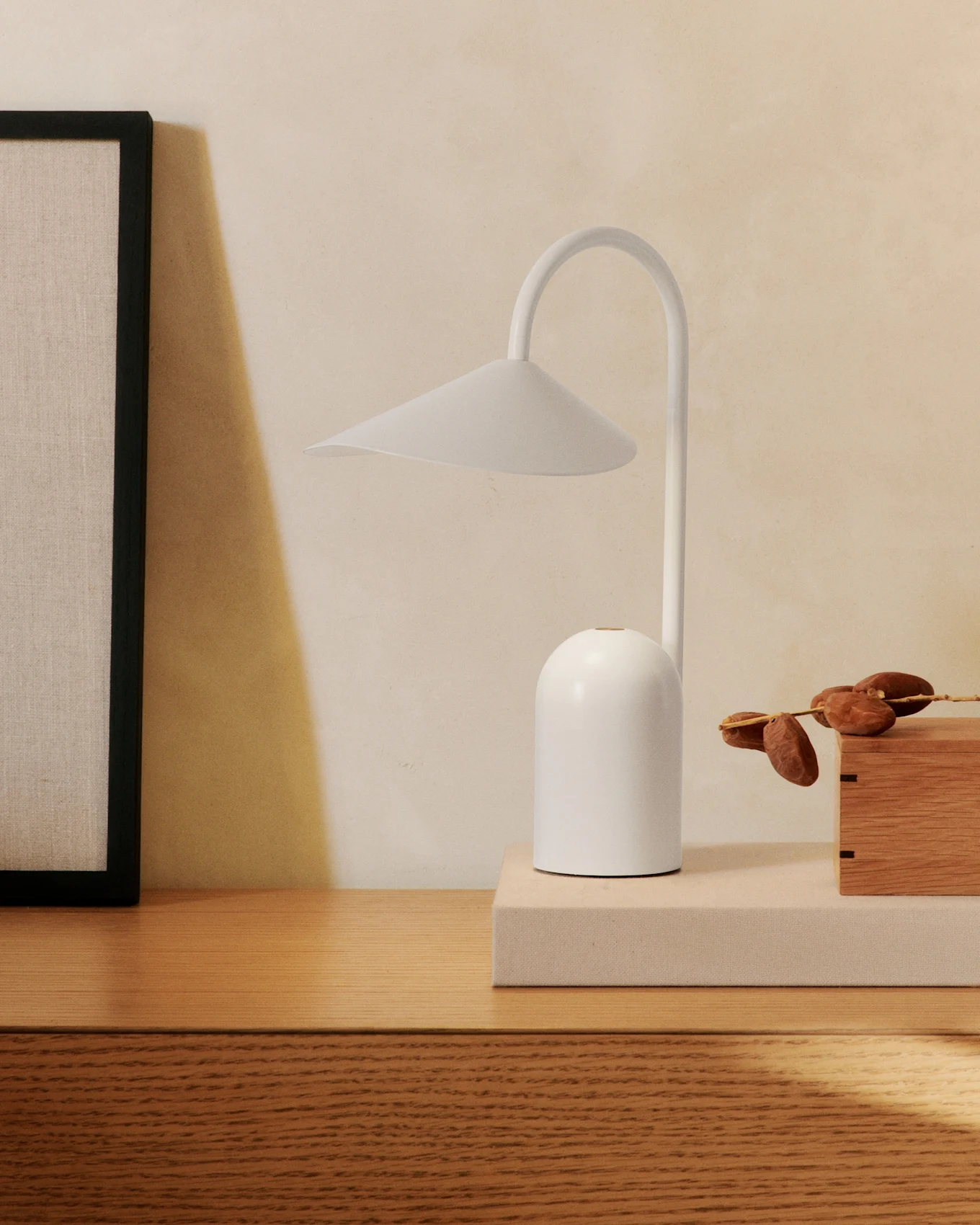 Eine minimalistische weiße Tischlampe mit gebogenem Arm auf einer Holzkommode, neben einem gerahmten Bild und einer Holzkiste mit Datteln.