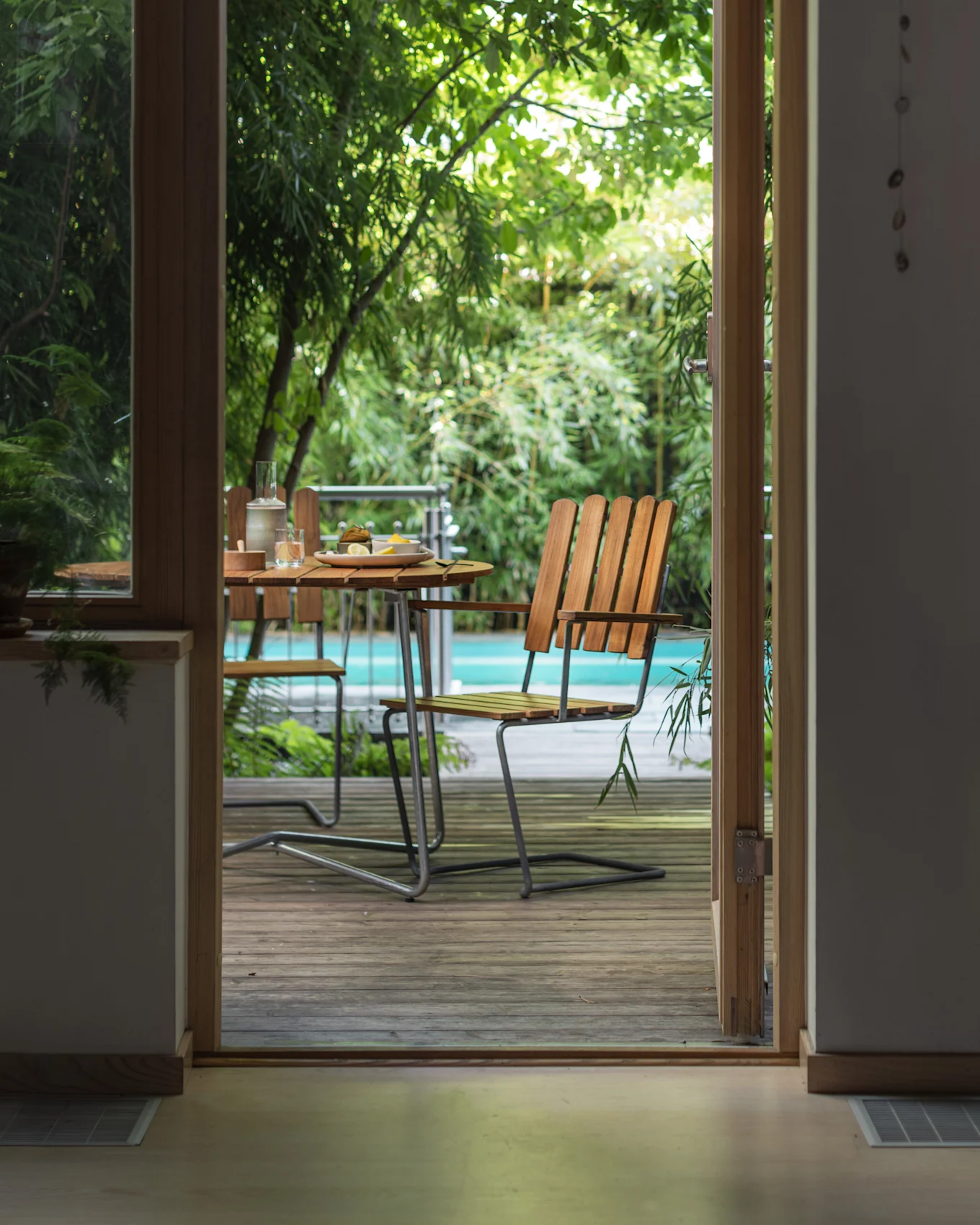 Blick durch eine offene Tür auf eine Außenterrasse mit einem Holztisch, Stühlen und einem Pool, umgeben von grünen Pflanzen.