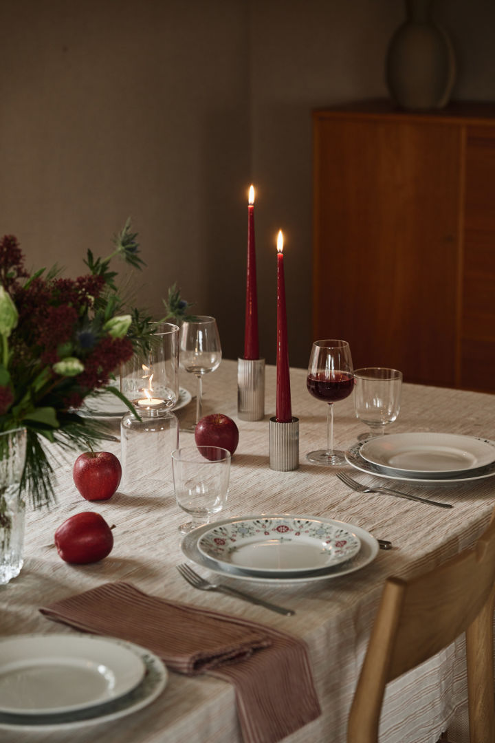 Rote Weihnachtskerzen stehen auf einem einfach gedeckten Weihnachtstisch mit gemusterten Tellern und roten Akzenten. 