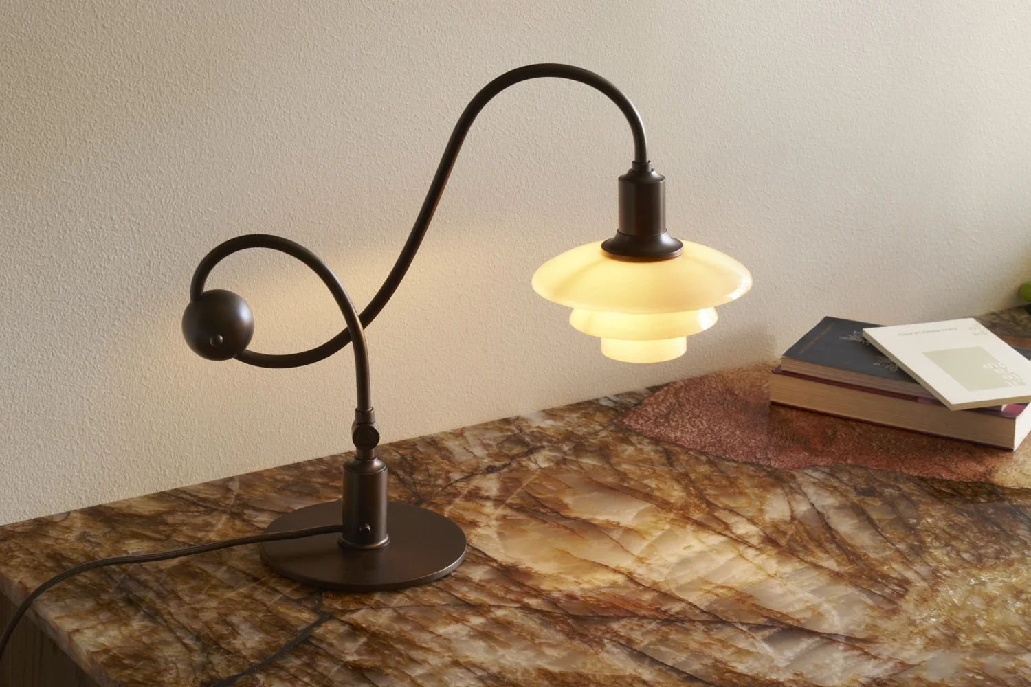 Dunkle Metall-Tischlampe mit gebogenem Stiel und gestuftem, leuchtendem weißem Schirm auf einer marmorierten Oberfläche mit Büchern.
