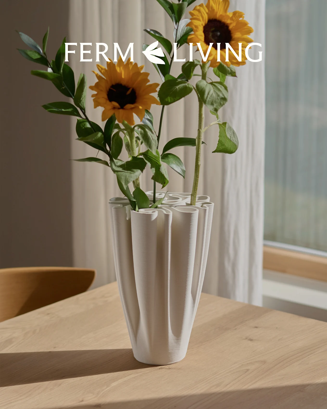 Sonnenblumen in einer weißen, gerippten Wellenvase auf einem Holztisch. Ferm Living Logo.