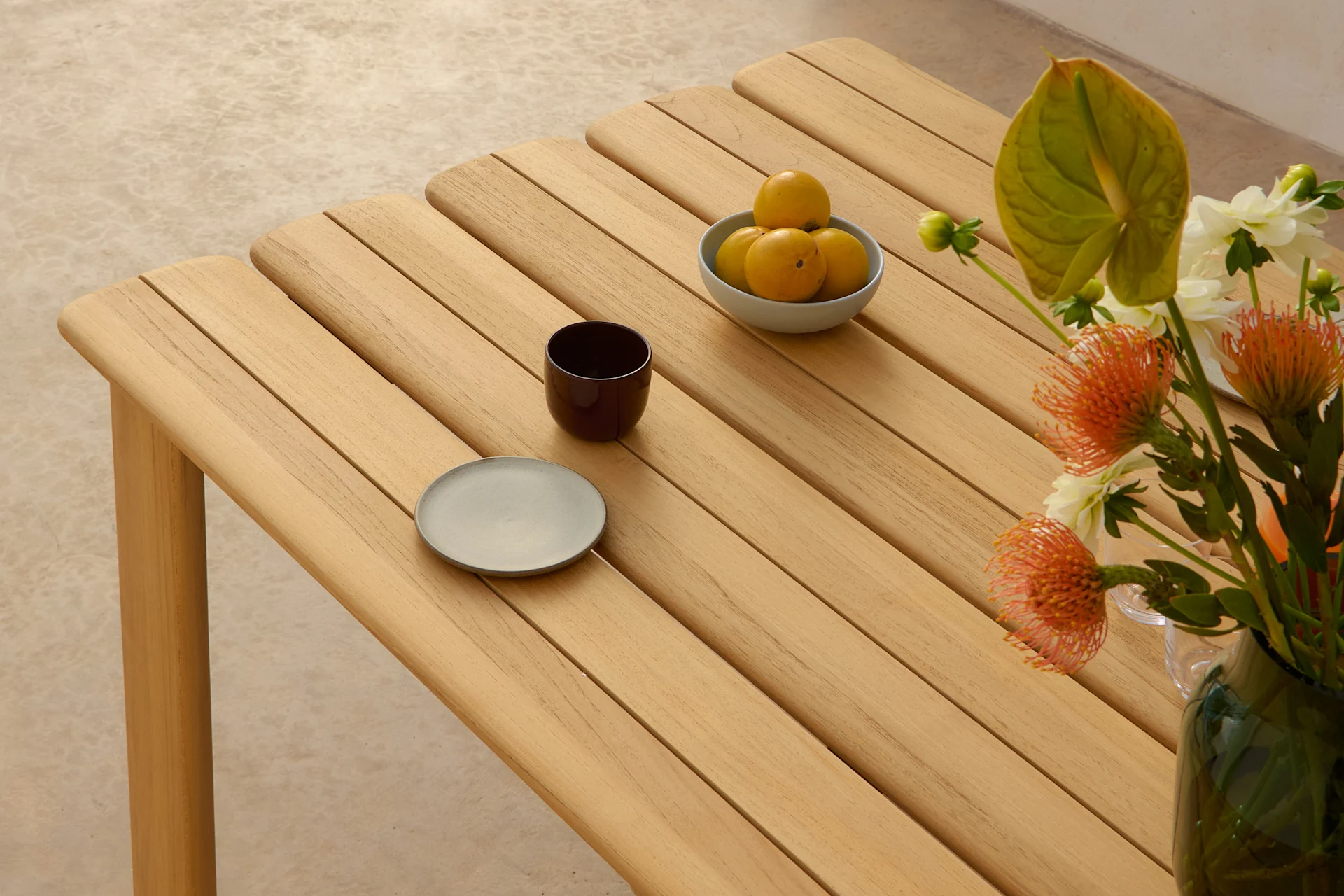 Ein heller Holztisch mit Lamellen, darauf eine Schale gelber Früchte, eine dunkle Tasse, ein grauer Teller und eine Vase mit orange-weißen Blumen.