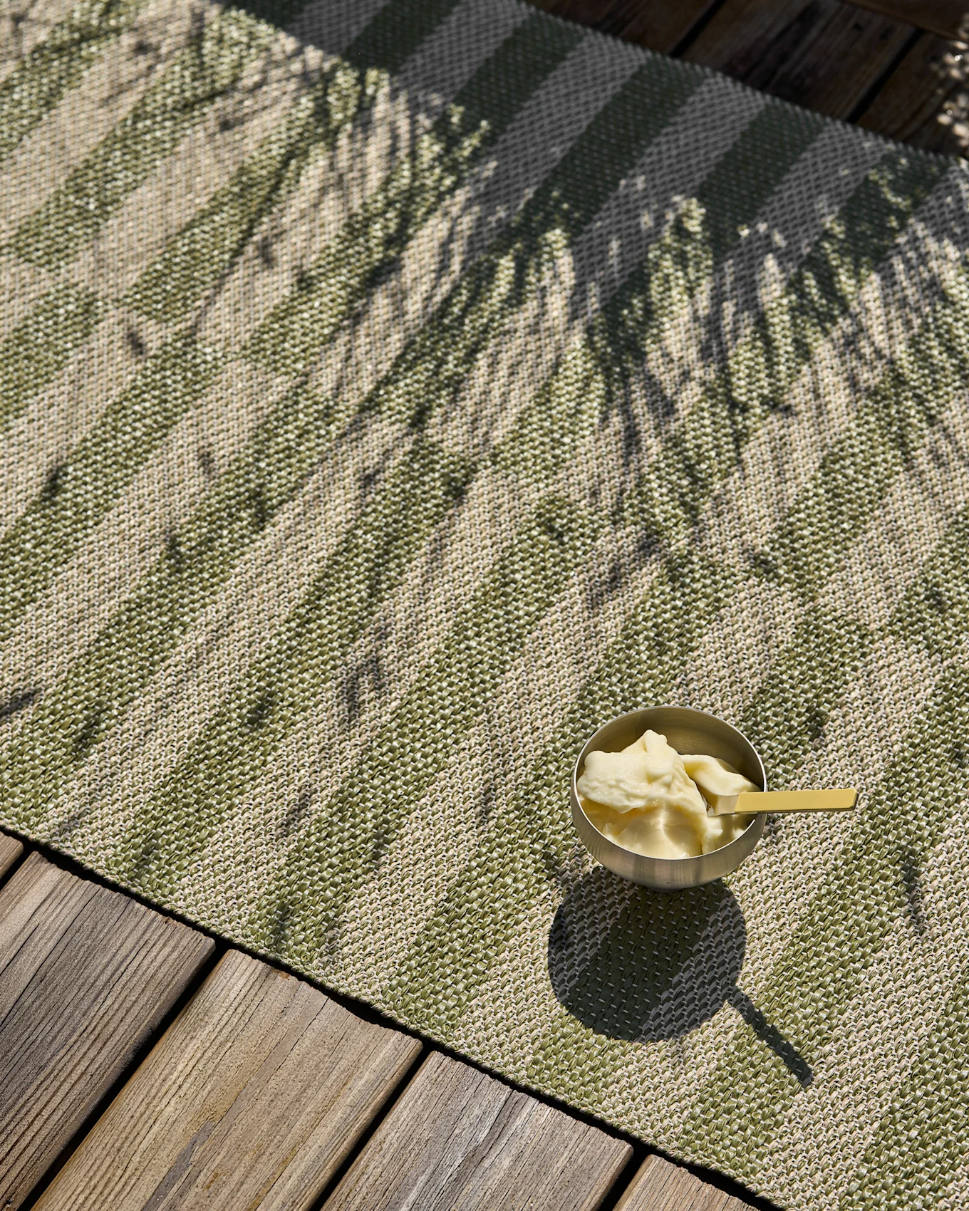 Grün und beige gestreifter Webteppich mit einer Eisschale und Palmenschatten auf einer Holzterrasse.
