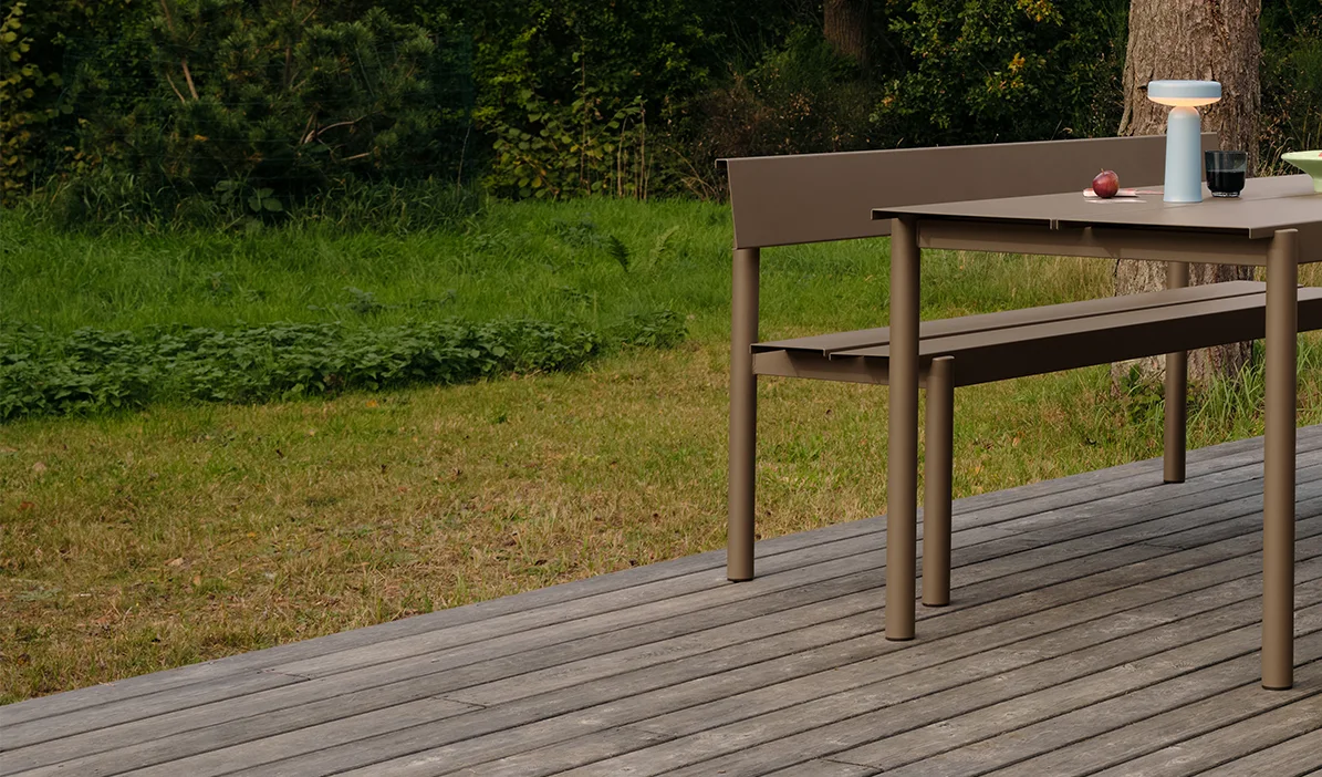 Moderner Esstisch und Bank in Taupe auf einer Holzterrasse, mit einer tragbaren Lampe und Äpfeln auf dem Tisch.