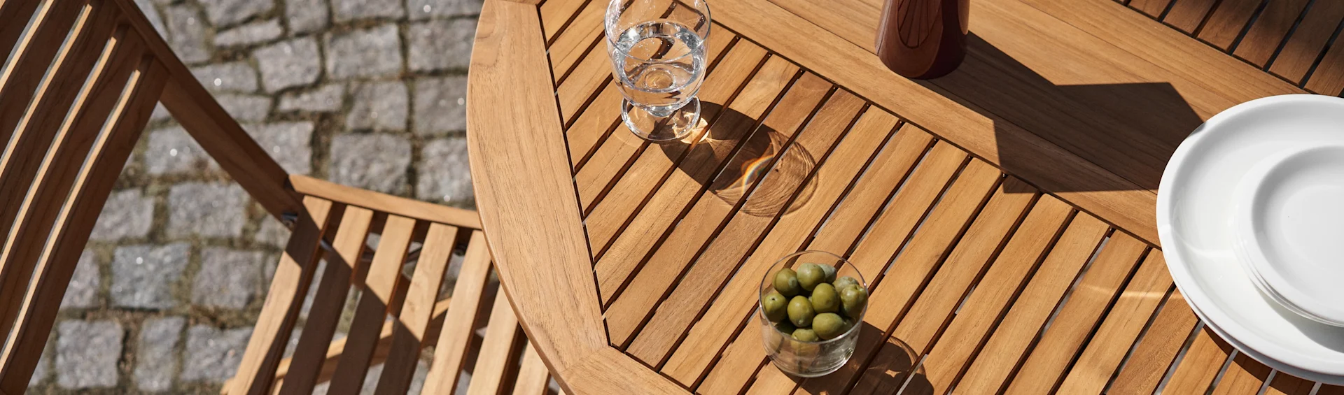 Aufnahme von oben auf einen Holzlamellen-Terrassentisch mit einem Glas Wasser, einer Schale grüner Oliven und weißen Tellern im Sonnenlicht.