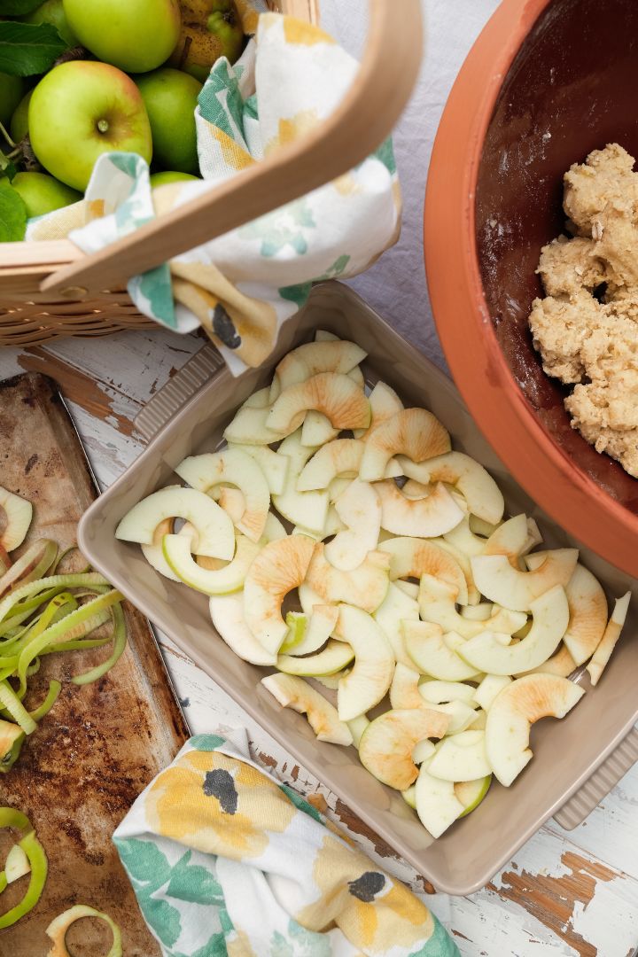 Das Bild zeigt die beigefarbene Flora-Auflaufform mit Henkel von Sagaform, hier gefüllt mit Apfelscheiben für einen Apfelkuchen. Daneben steht eine Knabstrup-Teigschüssel.