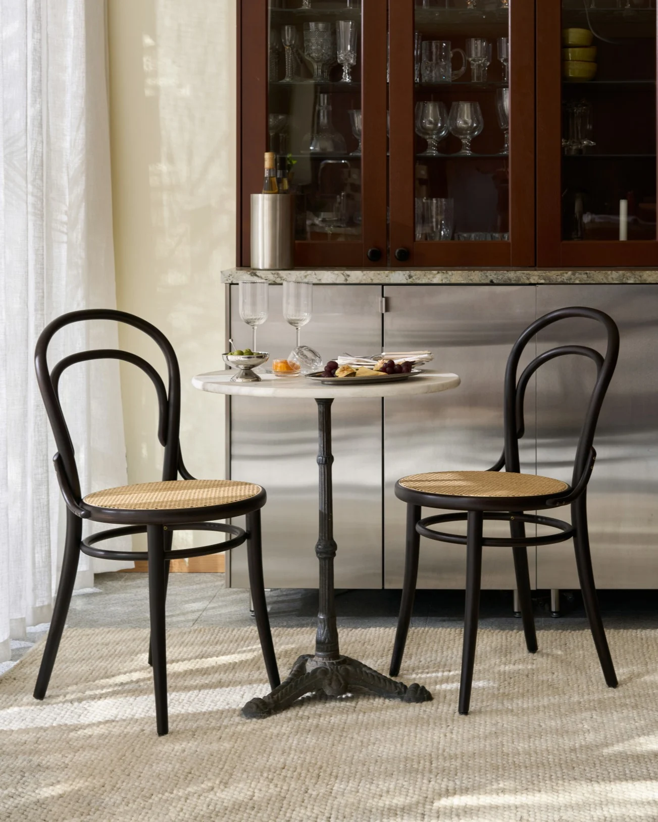 Ein Bistrotisch mit zwei dunklen Holz- und Korbstühlen, mit Snacks und Weingläsern gedeckt, vor einem Glasschrank.