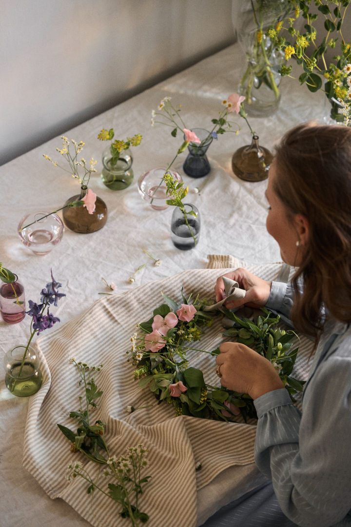 Eine Tradition zu Mittsommer in Schweden besteht darin, einen Blumenkranz aus selbstgepflückten Blumen zu basteln. Hier sehen Sie eine Frau an einem Tisch sitzen, während sie, umgeben von zahlreichen Mini-Midsummer-Vasen von Orrefors eine Blumenkrone kreiert.