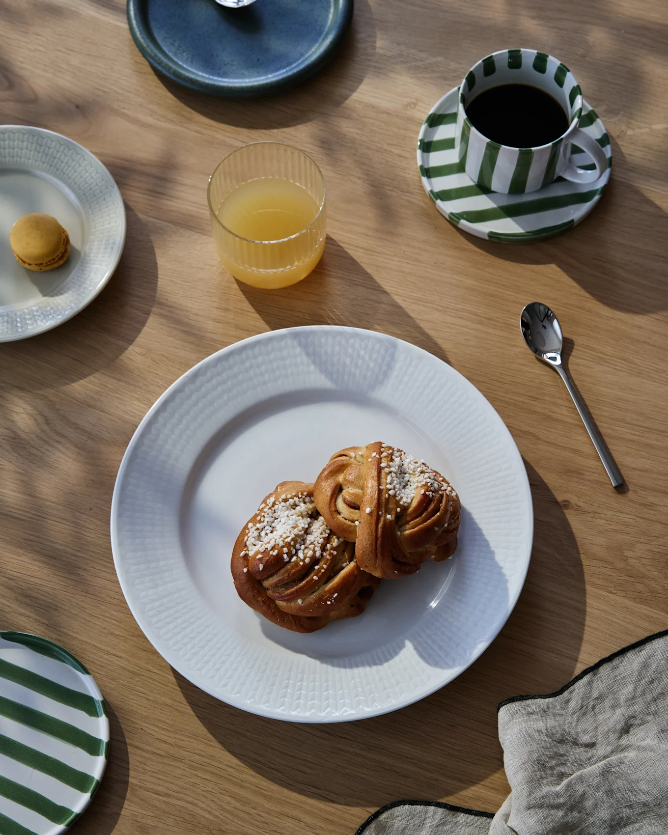 Ein Frühstück auf einem Holztisch mit zwei Zimtschnecken, Kaffee in einer gestreiften Tasse, Orangensaft und einem Macaron.