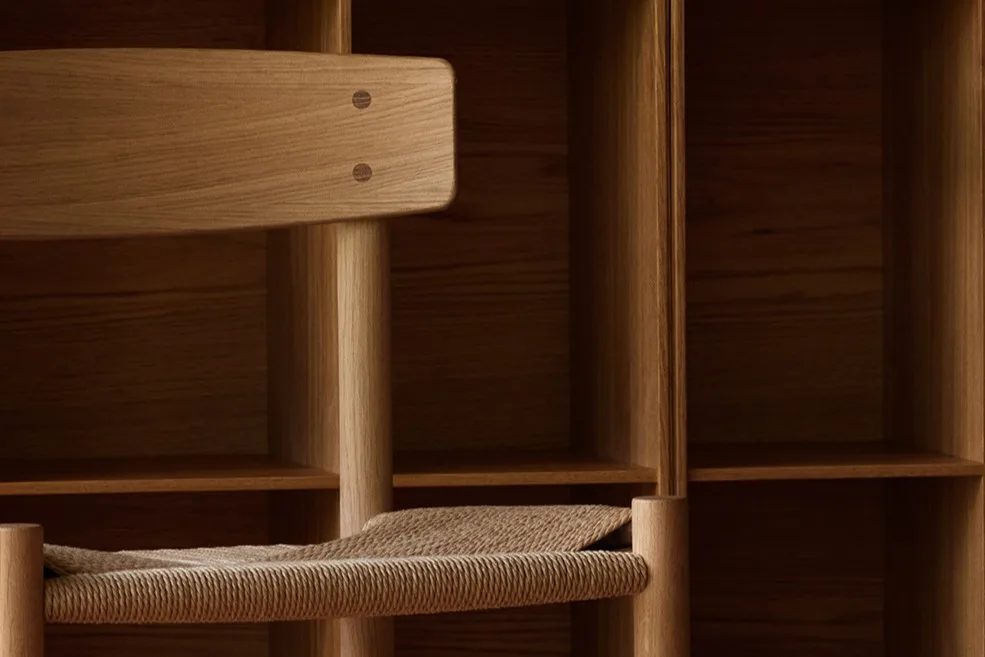 Ein heller Holzstuhl mit geflochtener Sitzfläche vor einer Wand aus quadratischen Holzregalen und einem gemusterten Holzboden.