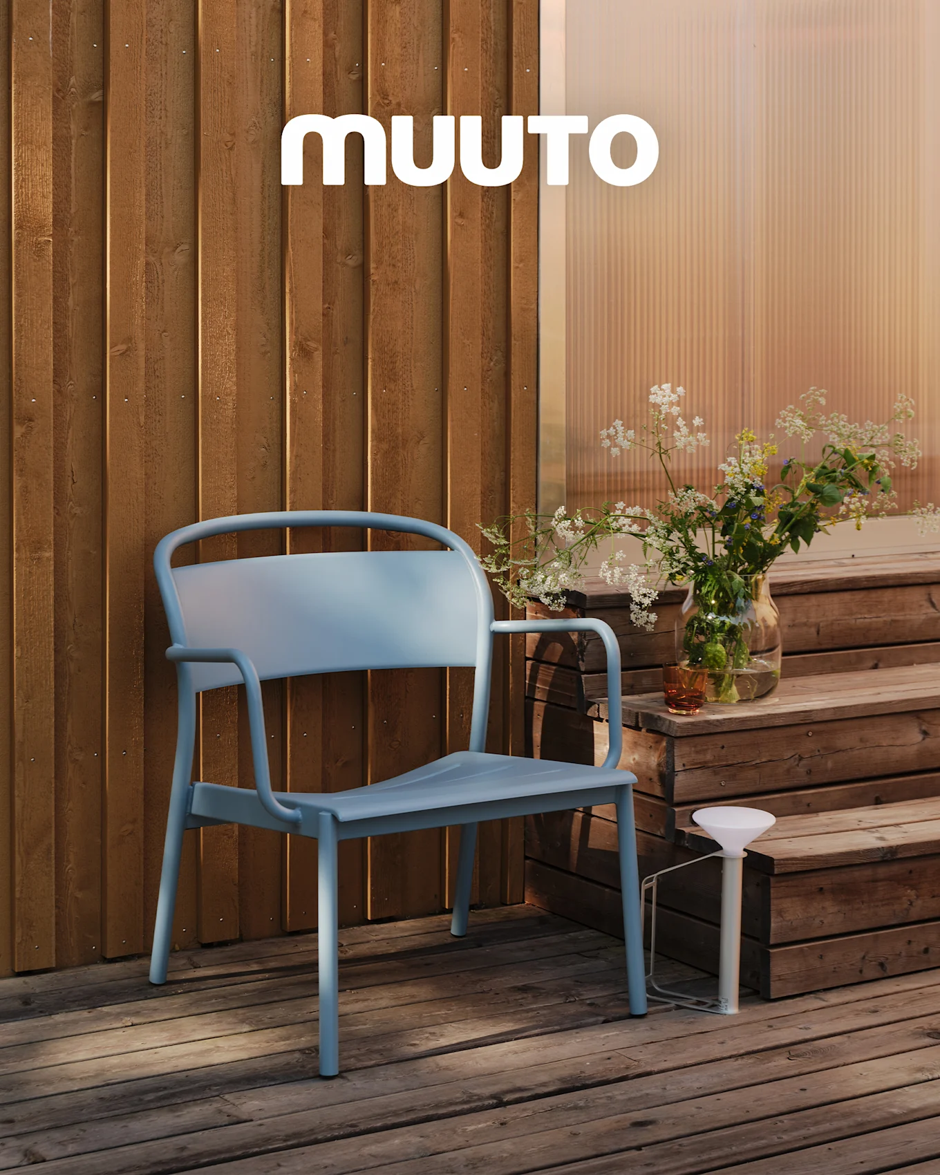 Ein hellblauer Outdoor-Stuhl auf einer Holzterrasse neben Stufen mit einer Vase Wildblumen und einer weißen tragbaren Lampe. MUUTO Logo.