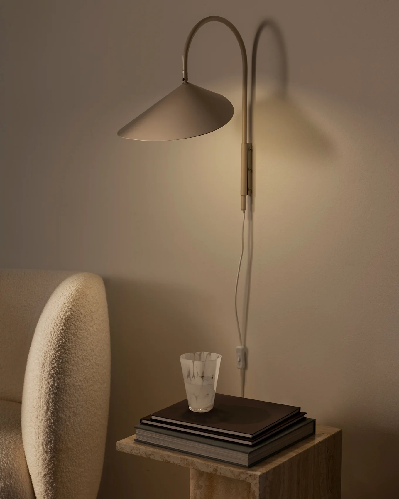 Warm beleuchtete Wandlampe mit konischem Schirm über einem Stapel Bücher und einem strukturierten Glas auf einem Steintisch, neben einem beigen Sessel.