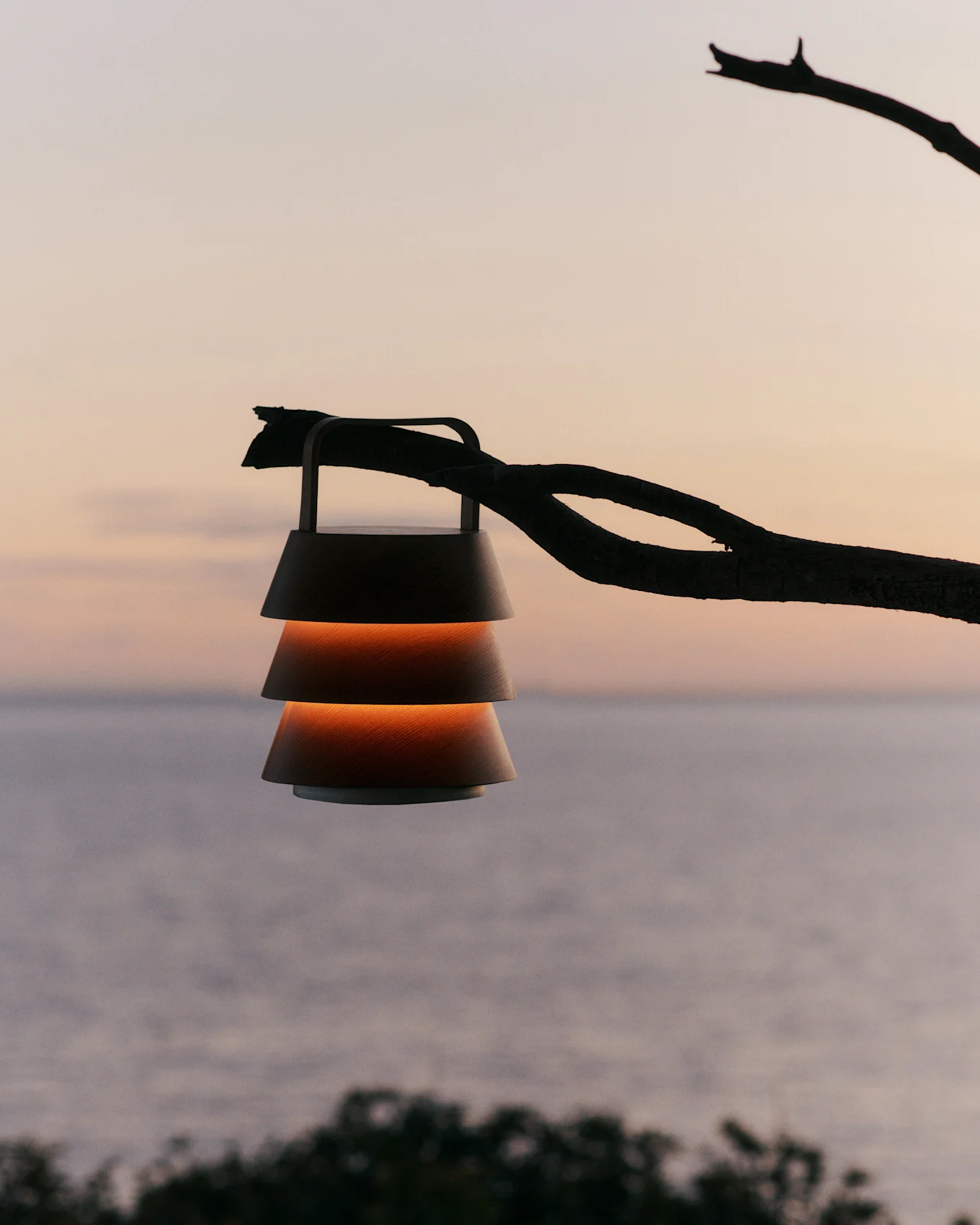 Laterne mit abgestuften Schirmen hängt an einem Ast vor einem Sonnenuntergangshimmel über Wasser.