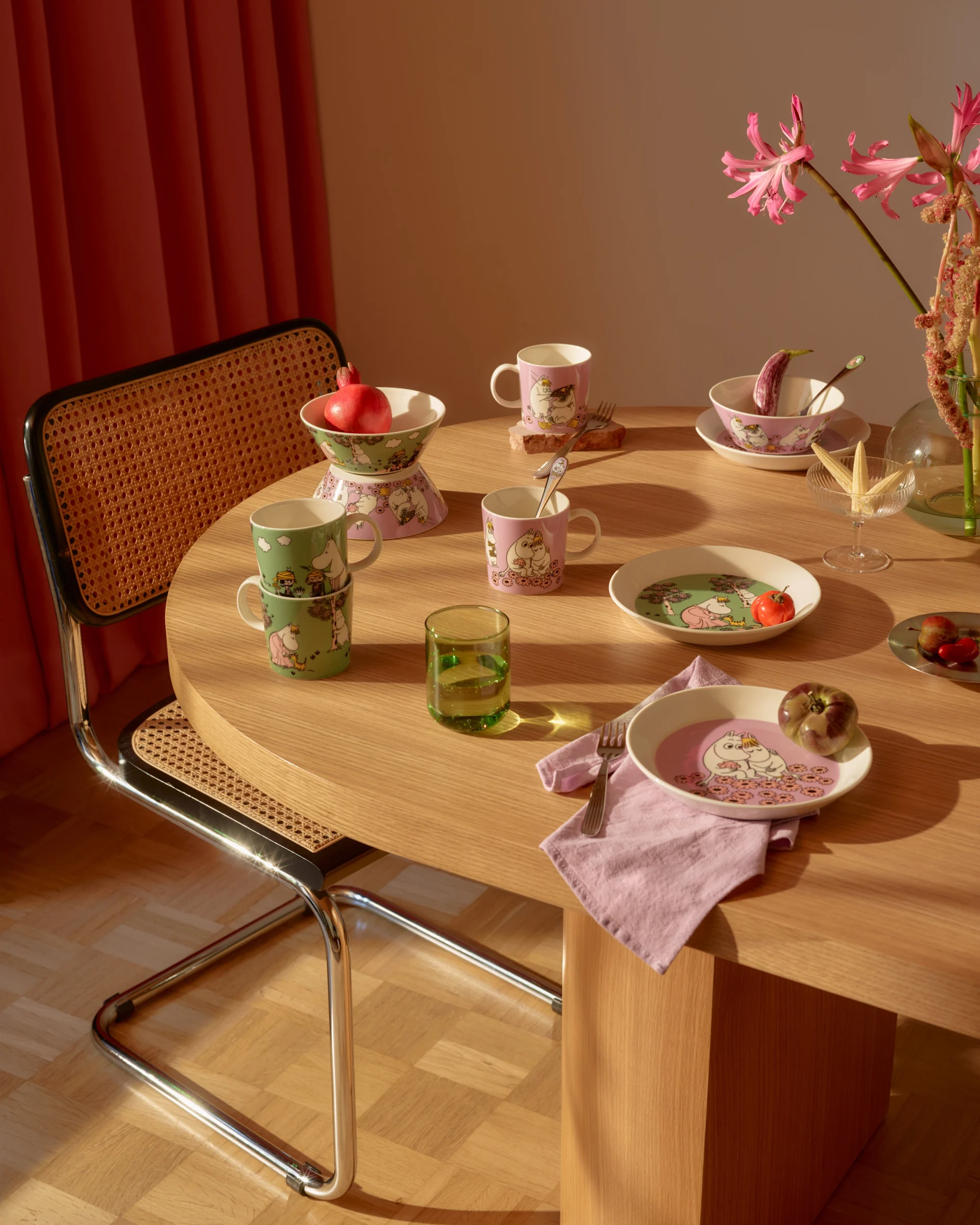Esstisch mit rosa und grünem Moomin-Geschirr, frischen Früchten, einem grünen Glas und rosa Blumen.