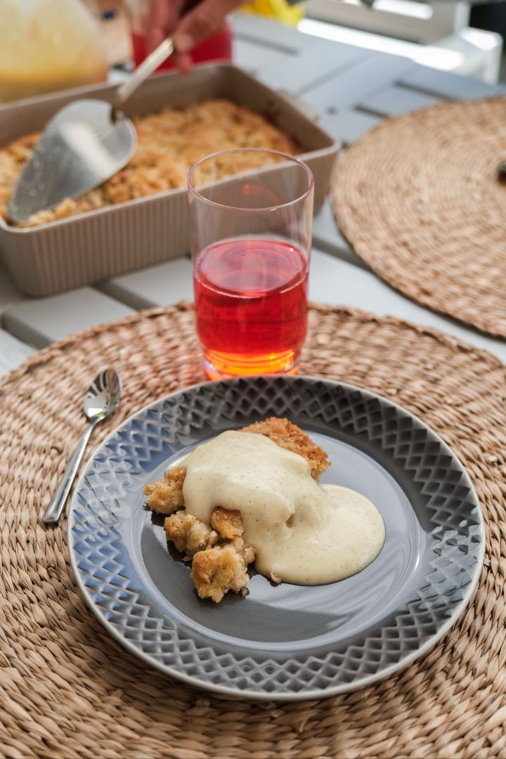 Das Bild zeigt einen Coffee More Teller in Grau mit Apfel Crumble und Vanillepudding darauf, sowie rotem Saft im Saga Trinkglas von Sagaform.