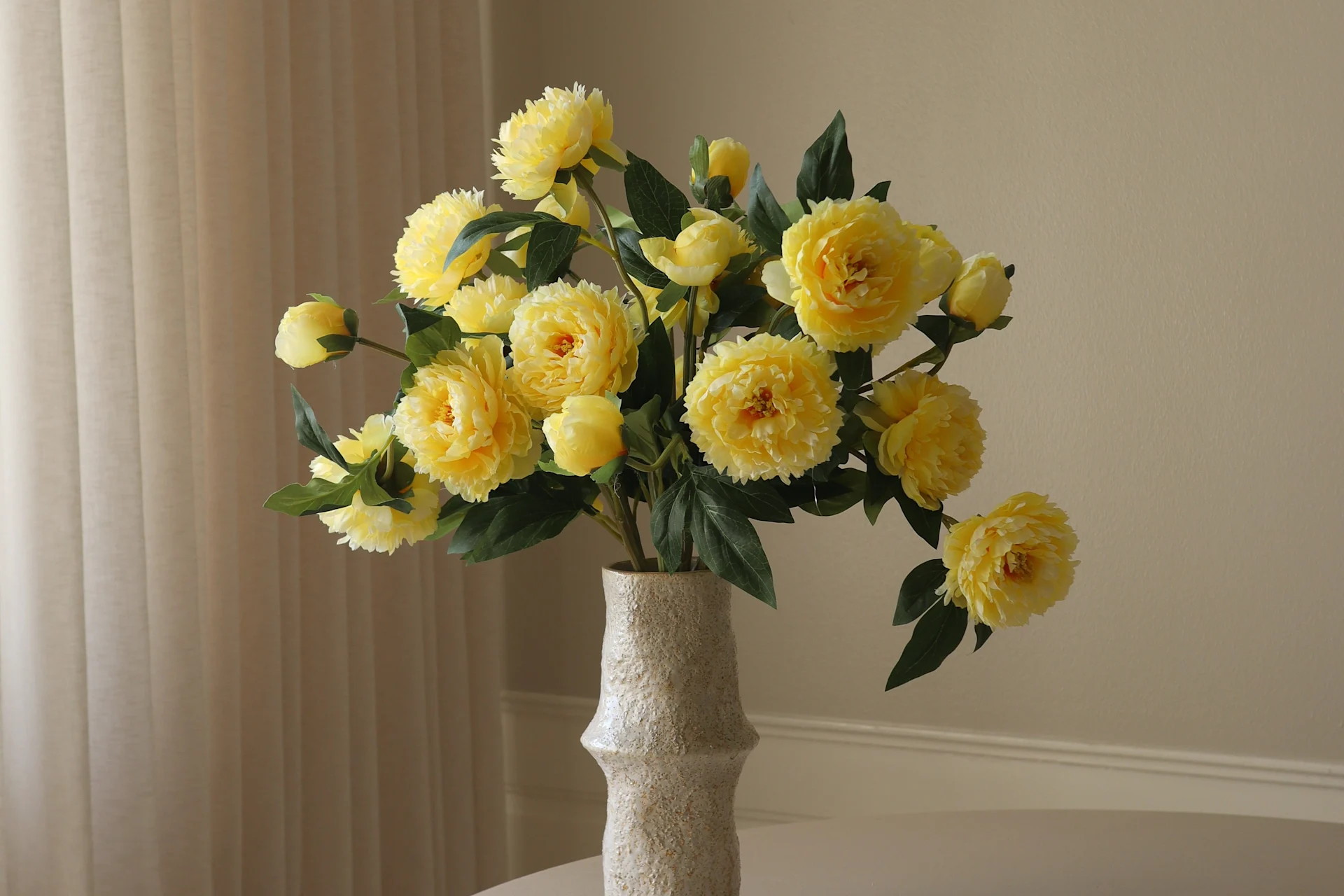 Eine helle, texturierte Vase enthält einen lebhaften Strauß gelber pfingstrosenartiger Blumen und grüner Blätter vor einem hellen Hintergrund.