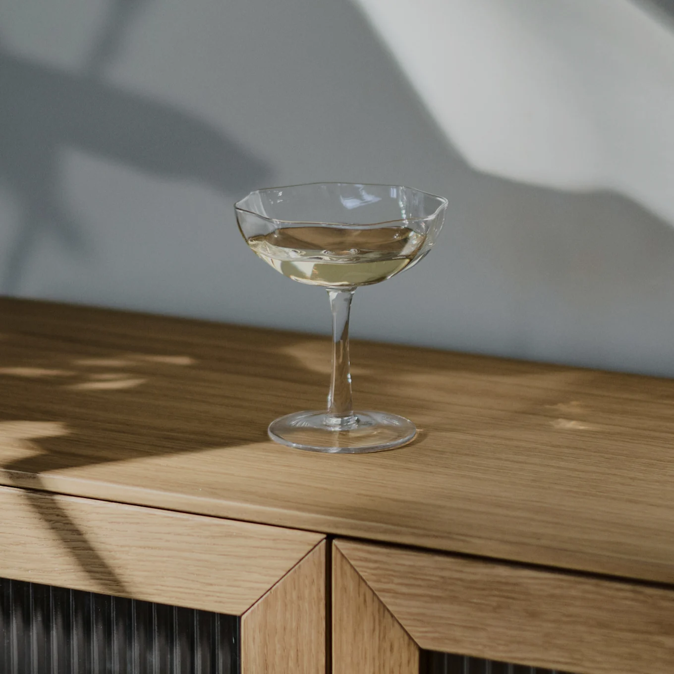 Ein klares Sektglas, gefüllt mit einem hellfarbigen Getränk, steht auf einem hellbraunen Holzschrank, mit Schatten an der Wand dahinter.