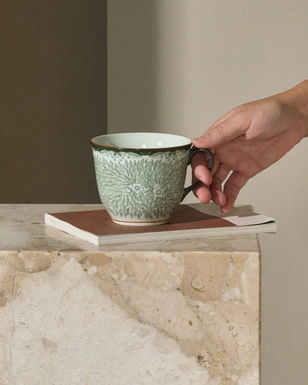 Person greift nach einer grün gemusterten Tasse auf einem Marmor-Beistelltisch neben einem Sofa.