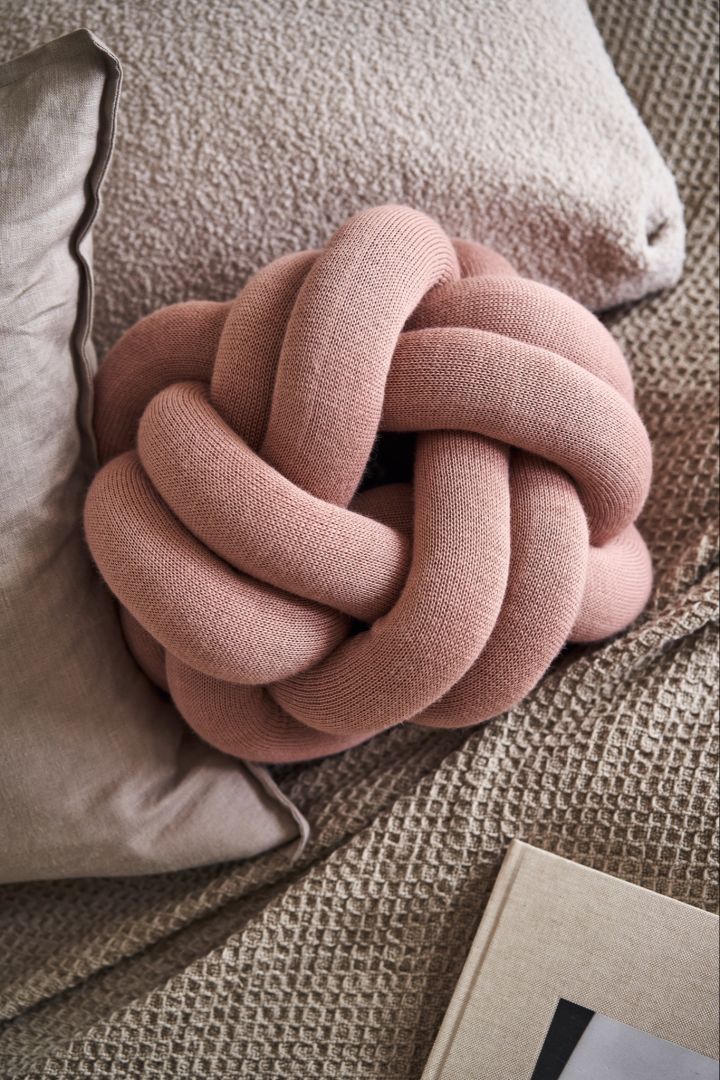 Das kleine Knotenkissen in Rosa liegt auf einem Bett mit beiger und grauer Bettwäsche. 