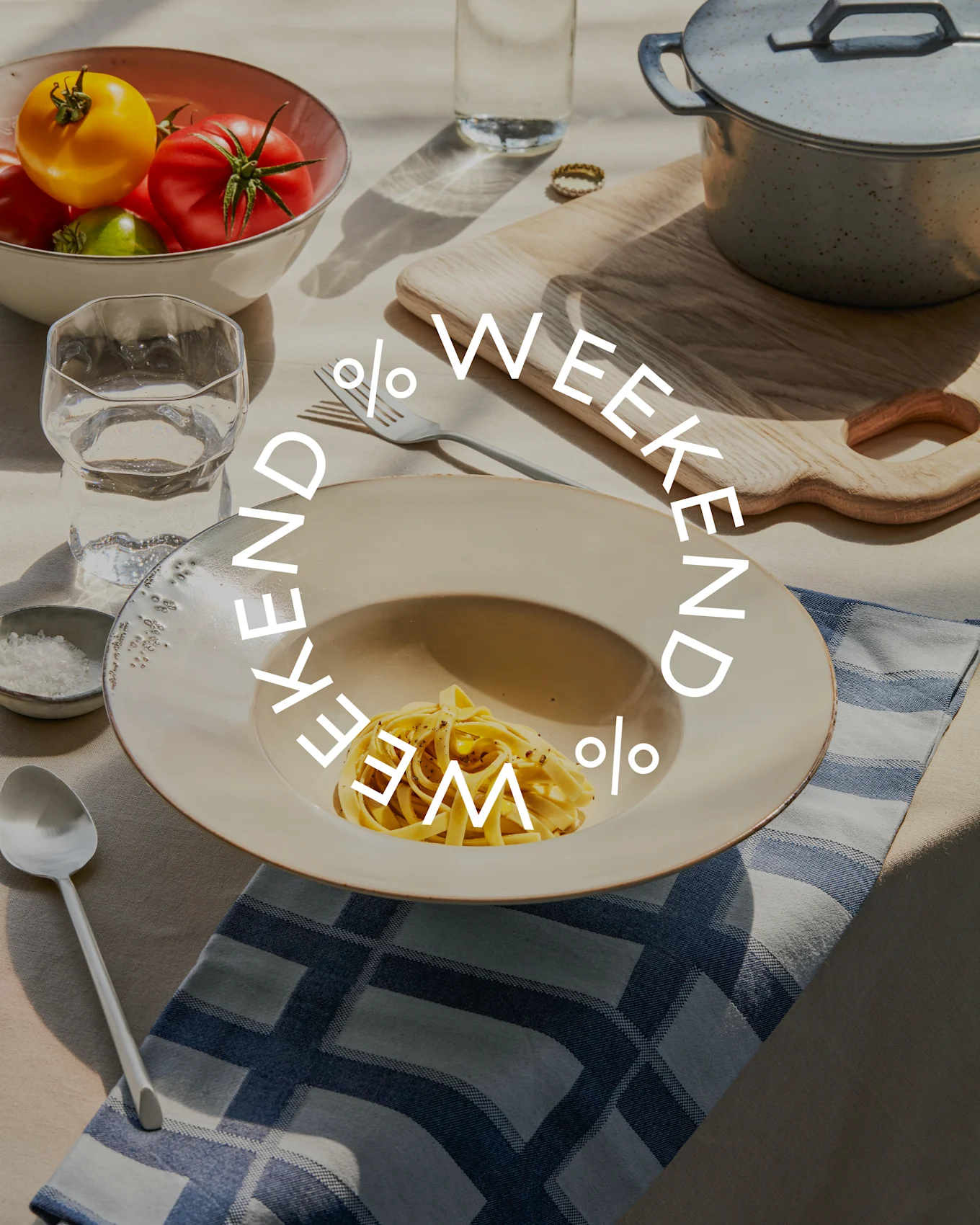 Tisch mit Pasta, einer Schale frischer Tomaten, Wasserglas und Kochtopf. Der Text 'WEEKEND % WEEKEND %' ist überlagert.