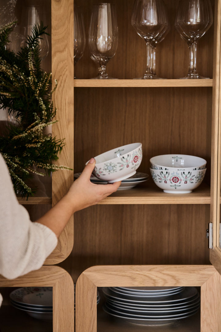 Eine Hand nimmt eine gemusterte Weihnachtsschale aus einem Schrank, um sie auf einen weihnachtlich gedeckten Tisch zu stellen. 