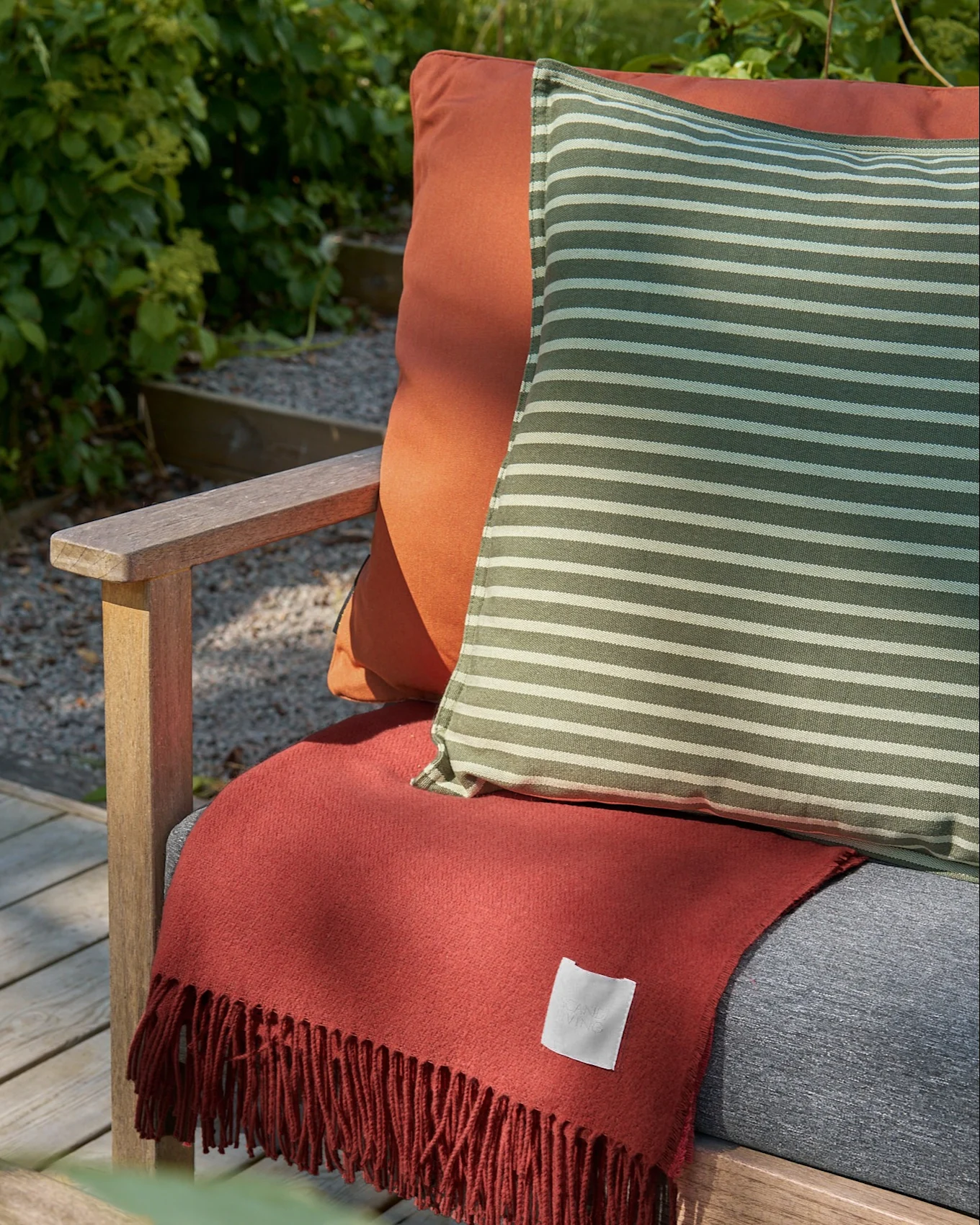 Outdoor-Holzstuhl mit orangefarbenem Kissen, einem grün-weiß gestreiften Kissen und einer terrakottafarbenen Fransendecke.