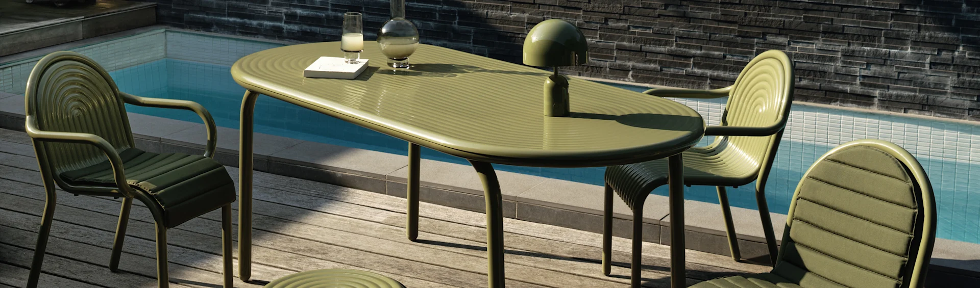 Grüner gerippter Outdoor-Esstisch und Stühle auf einer Holzterrasse an einem Pool. Eine Lampe, Karaffe, ein Glas und ein Buch stehen auf dem Tisch.