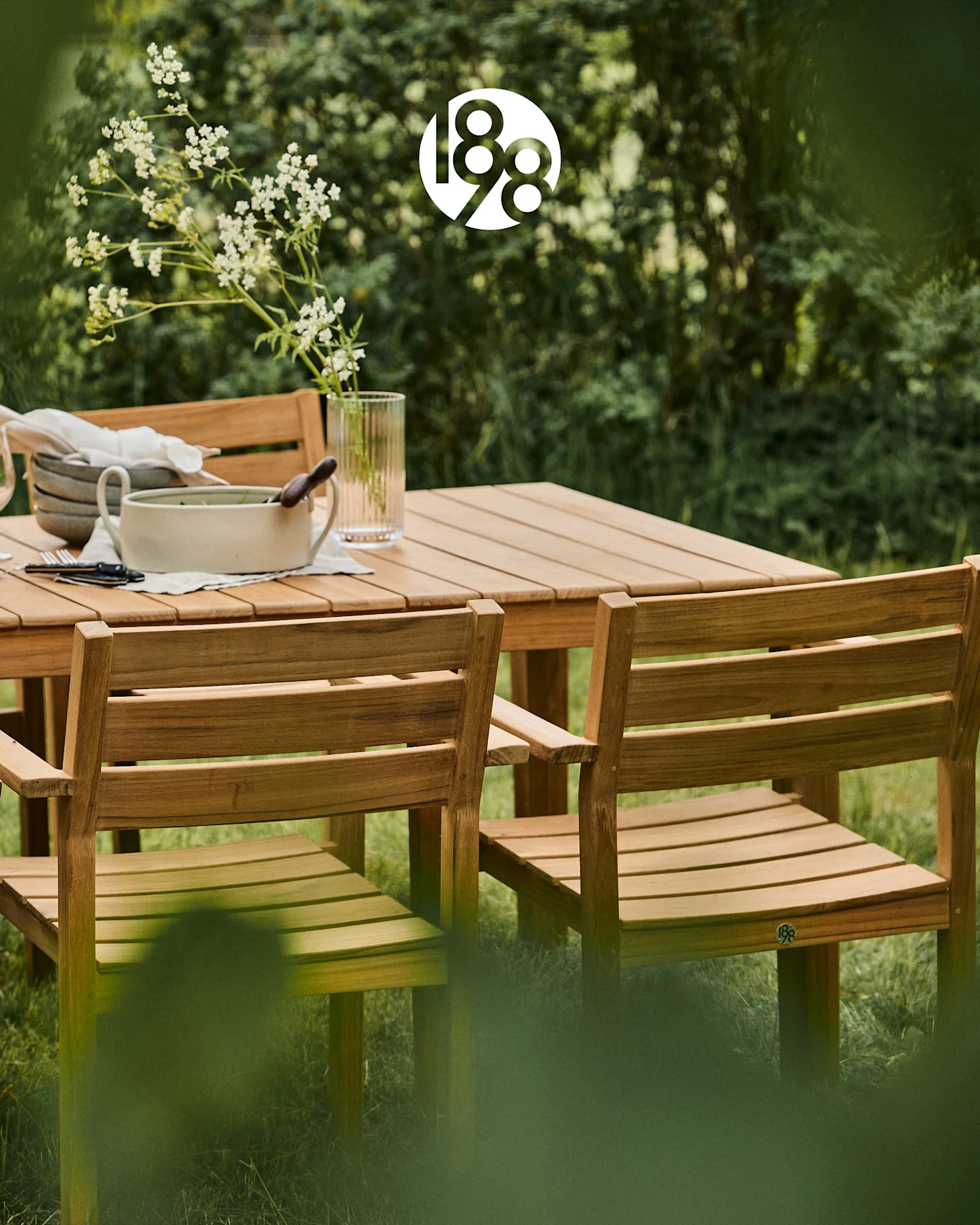 Hölzernes Outdoor-Esszimmer-Set mit Tisch und Stühlen in einem üppig grünen Garten, mit weißen Blumen und Serviergeschirr.
