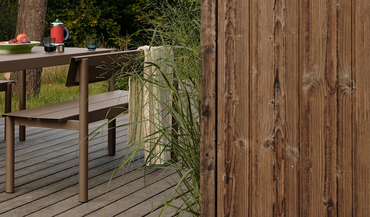 Brauner Metall-Gartentisch und Bank auf einer Holzterrasse mit Obst und Getränken, neben hohem Gras und einem Holzzaun.